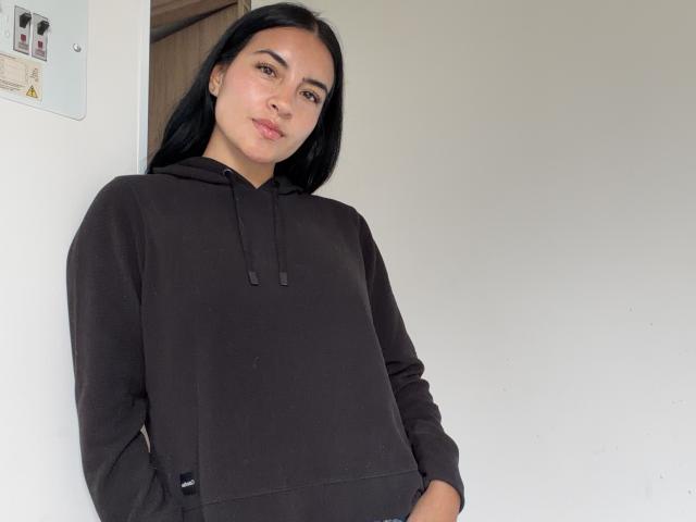 EmilyJensenn stands against a white wall, wearing a black, long-sleeve hoodie with a drawstring. She has long, straight black hair, and her brown eyes are slightly downcast. Her light brown skin contrasts with the dark hoodie. Her hands are in the hoodie pockets. The left wall has a small, white electrical panel with a few stickers. Natural light highlights her relaxed, slightly smiling expression. The composition is simple and focused on EmilyJensenn.
