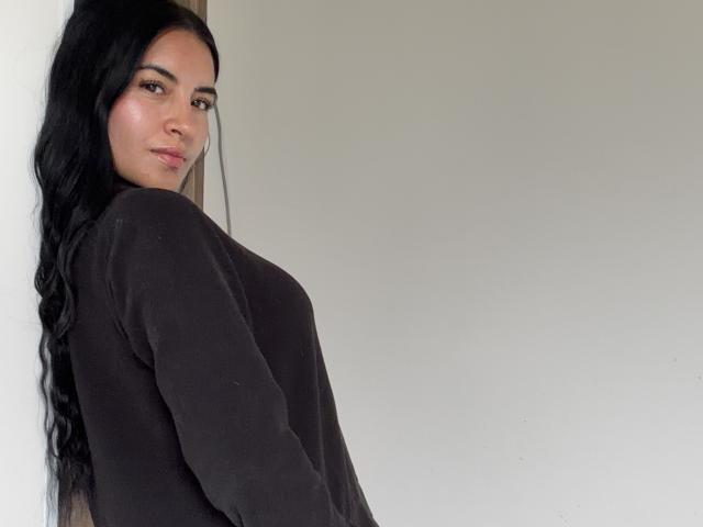 EmilyJensenn, a woman with long black wavy hair, stands against a white wall. She has a medium build with noticeable curves and wears a loose black sweater. Her brown eyes are slightly downcast, and she has a subtle, confident expression. The light is soft, highlighting her smooth, slightly shiny skin. The image is a straightforward, unfiltered portrait. Her posture is slightly turned to the side, emphasizing her profile. The composition is simple, with a focus on EmilyJensenn's face and upper body.