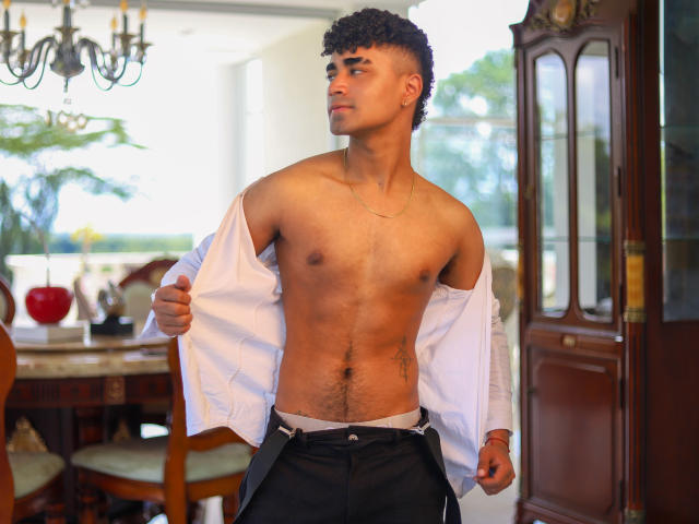 Jacobb69 with curly black hair, shirtless, white shirt open, revealing a toned, hairy chest and a visible white waistband, standing in a room with wooden furniture.