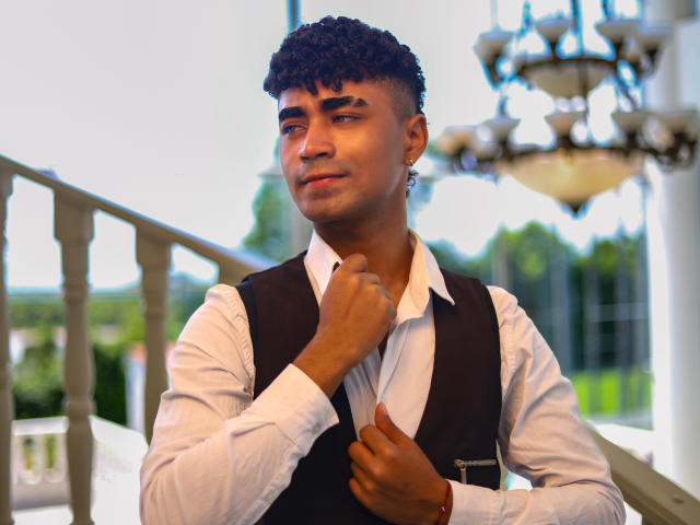 Jacobb69 with curly black hair, brown skin, and a slight smile, wearing a white shirt and black vest, holding his collar, standing by a balcony with a chandelier.
