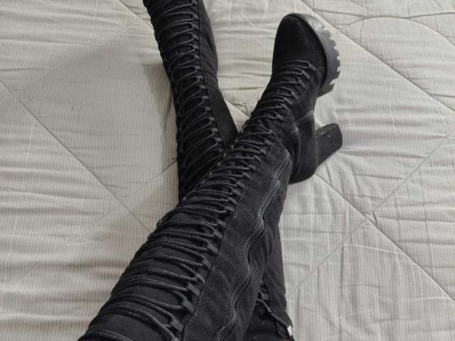 VittoriaI's black lace-up boots with textured leather, standing with legs crossed, showing black soles on a quilted, white bed. Boots are tight, highlighting VittoriaI's thighs.
