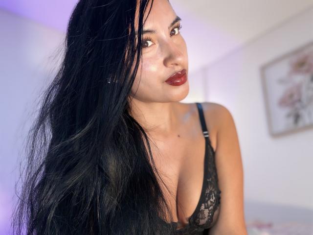 EmilyJensenn with long black hair, dark red lipstick, and black lace bra, showing her big tits, gazing at the camera with a slightly parted mouth.