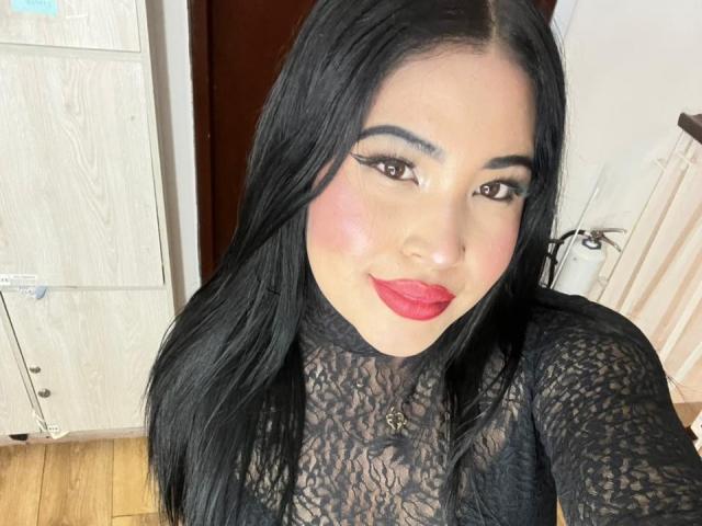 OliviGrace with long black hair, wearing a lace black top, has pink blushed cheeks, red lipstick, and a necklace. Her tits are slightly visible through the lace.