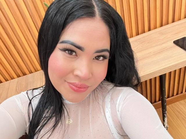OliviGrace with black hair, brown eyes, and pink lipstick, wearing a white, sheer top with small sparkles, and a heart necklace, taking a selfie in a wooden setting.