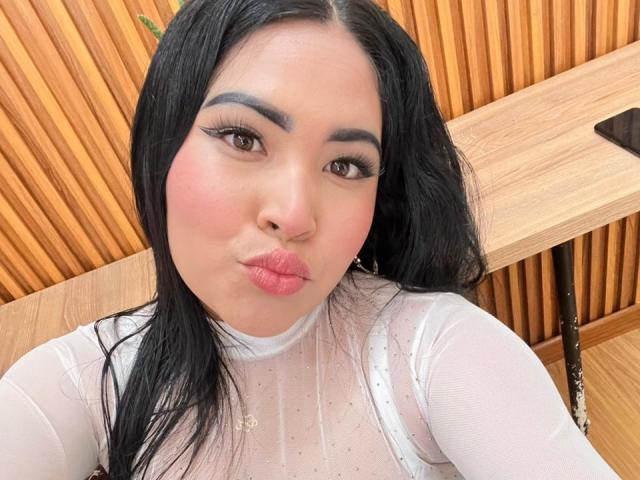 OliviGrace with puffy pink lips, black hair, and white sheer top, showing off her tits with subtle polka dots. Wooden background. She's sucking on her lips.