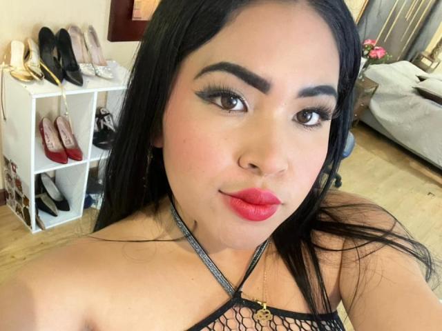 OliviGrace with long black hair, thick eyebrows, red lipstick, and a black fishnet top, showing off her tits, wearing a chain necklace, in a bedroom.