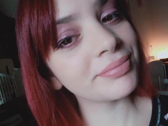 ValeryLoveLovely mit roten Haaren, rosa Lidschatten und glänzenden rosa Lippen, lächelt leicht mit einem süßen, dezenten Schmollmund. Ihre Augen sind halb geöffnet, und ihre Haut ist blass..