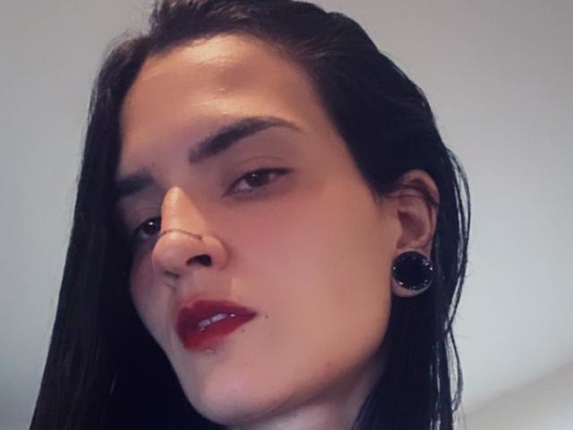 EmmaHerby has dark wet hair, red lipstick, black ear gauges, and a nose ring, looking sideways with a slight smirk. Her skin is light with subtle makeup.