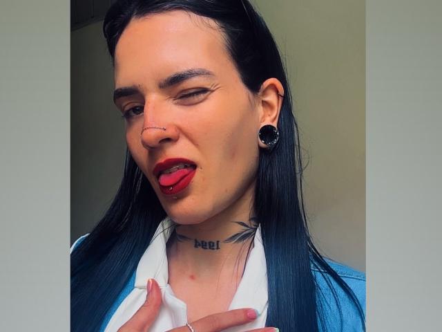 EmmaHerby with black hair, nose ring, red lipstick, tongue out, blue jacket, white shirt, black ear gauges, tattoo under chin, and a tattooed neck, winking.
