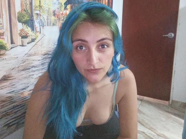 KarolLove69 with blue-green hair, medium tits, brown eyes, wearing a green spaghetti strap top, looking at camera, in a room with a wooden door.