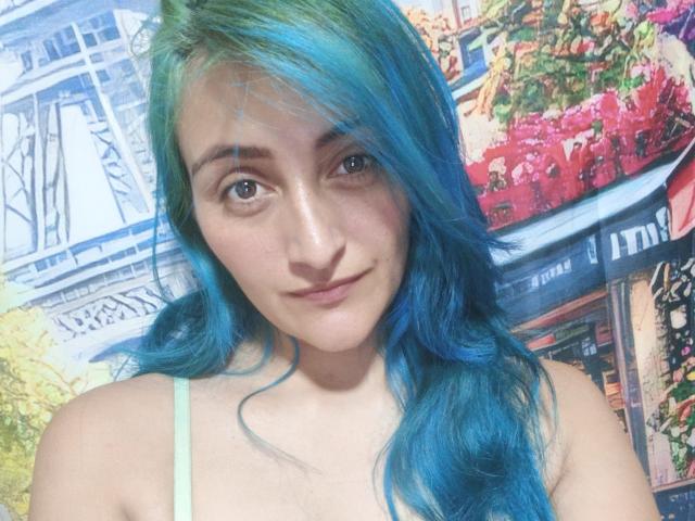 KarolLove69 with blue-green hair, light skin, small tits, wearing a green strap, smiling softly, blue eyes, wavy hair, standing in front of a colorful, vibrant background.