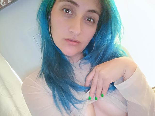 KarolLove69 with blue hair, green nails, wearing a sheer top, showing her tits, looking at the camera, hand on chest, soft expression, light skin, hoop earrings.