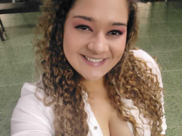 NataliaWendys with curly brown hair, pink eyeshadow, and white shirt, smiling at camera, showing large boobs, green tiled floor in background.