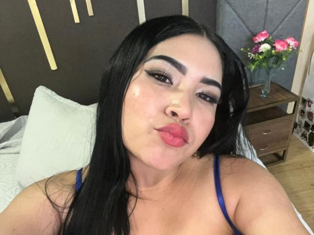 OliviGrace tem cabelo preto, alças finas azuis, pele brilhante e lábios vermelhos cheios, fazendo uma carinha de beijo com sobrancelhas escuras e grossas e cílios longos..