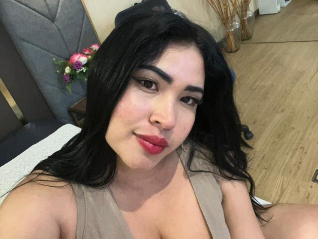 OliviGrace heeft lang zwart haar, donkere wenkbrauwen en roze lippenstift, draagt een beige top, waarbij haar borsten zichtbaar zijn, en zit met een zelfverzekerde glimlach in een houten kamer..