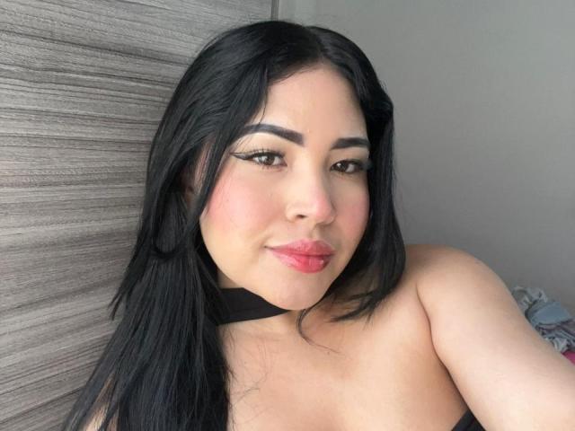 OliviGrace has black hair, pink lips, and thick black eyebrows. She's wearing a black choker and has a slight smile, showing her puffy ass in the background.