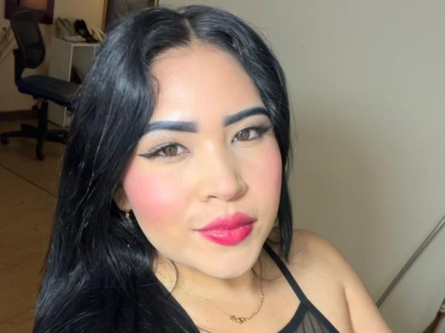 OliviGrace with black hair, blue eyebrows, pink blush, red lipstick, gold necklace, and black halter top, smiling at the camera, showing off her cute face and sexy look.