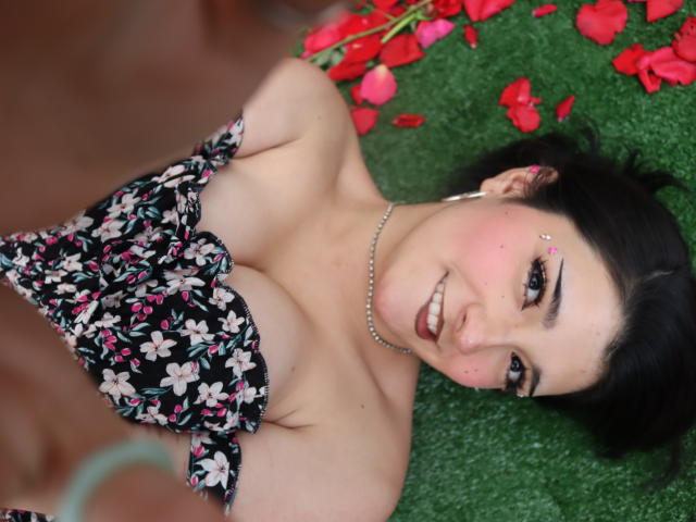 SweetEescape has dark hair, a nose piercing, and is smiling at the camera with medium tits in a black floral dress, lying on green grass with red petals.