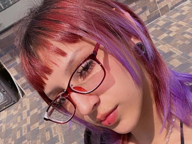 KatsuraRen with red and purple dyed hair, wearing glasses and a lip piercing, has black ear gauges and a neutral expression. Sunlight highlights her face.