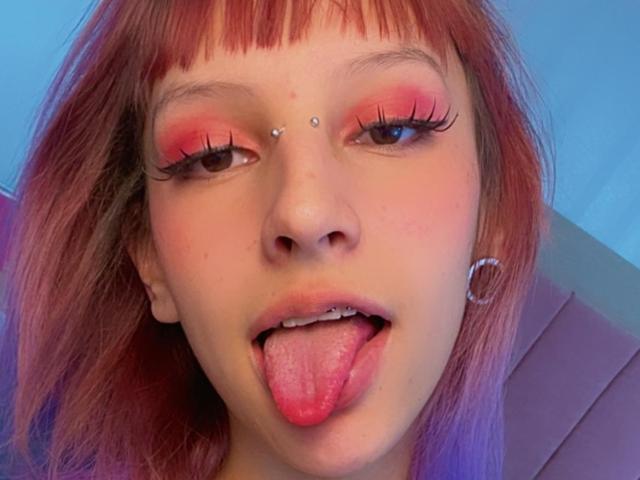 KatsuraRen has red hair, pink eyelid makeup, a nose piercing, and a tongue out, showing a silver hoop earring, with a cute and playful expression.
