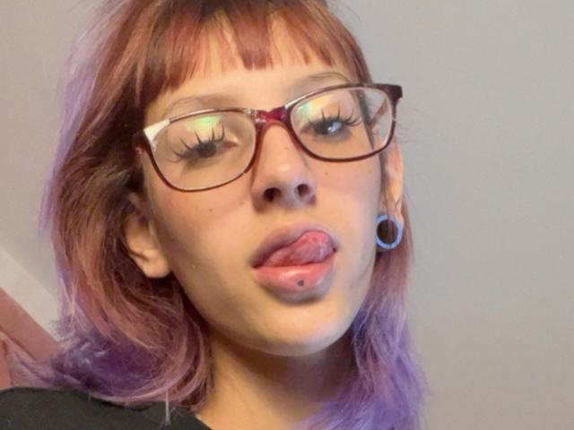 Close-up of KatsuraRen with pink-purple hair, wearing large red-framed glasses, sticking out tongue with a lip piercing. KatsuraRen has blue ear stretchers, long eyelashes, and a light skin tone. The hair is straight with bangs, and the expression is playful. The background is plain and out of focus, emphasizing KatsuraRen's face. The image has a casual, selfie-style composition. The focus is on KatsuraRen's facial features and accessories.