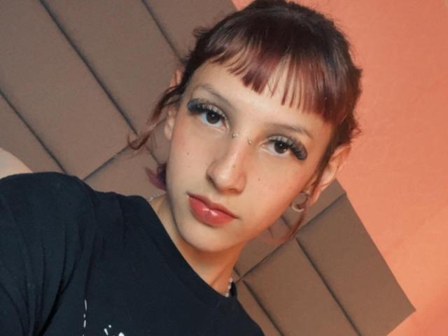 KatsuraRen has red-brown hair, a nose piercing, and black eyeliner. She's wearing a black shirt, with a neutral expression, and has a small nose stud.