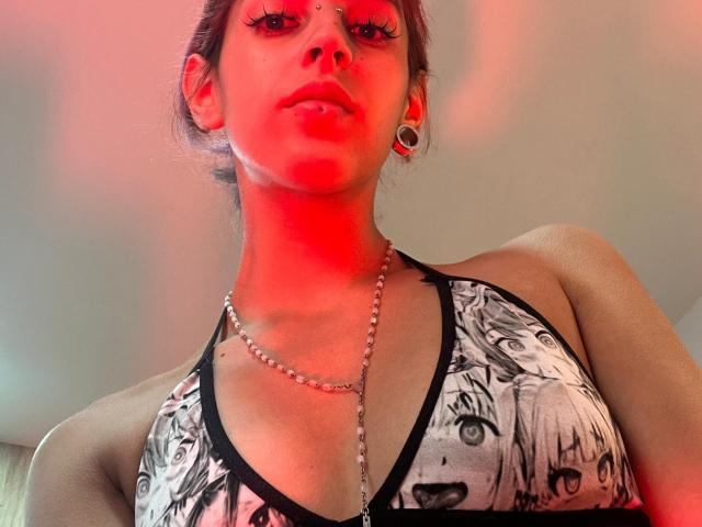 KatsuraRen with red lighting, wearing a black and white anime print top, chain necklace, and earrings, looking down at the camera, showing her tits and nose ring.