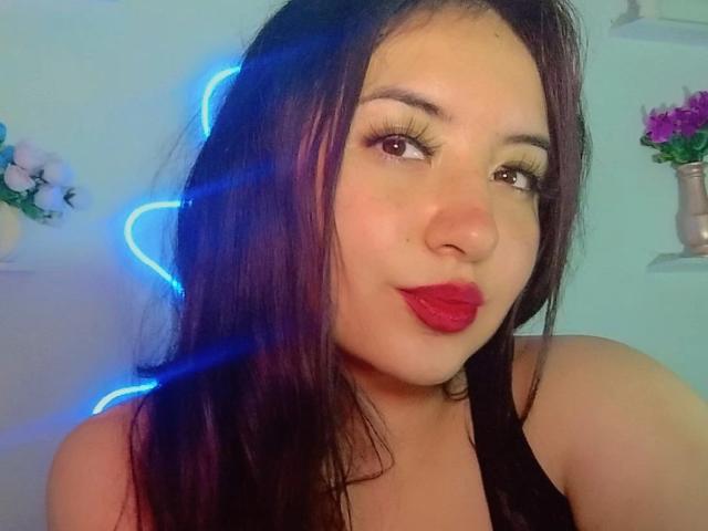 Susan Monroy, con cabello oscuro, luz neón azul, lápiz labial rojo y pestañas largas, mostrando su linda cara y una blusa negra. Pechos parcialmente visibles. Sonrisa suave..