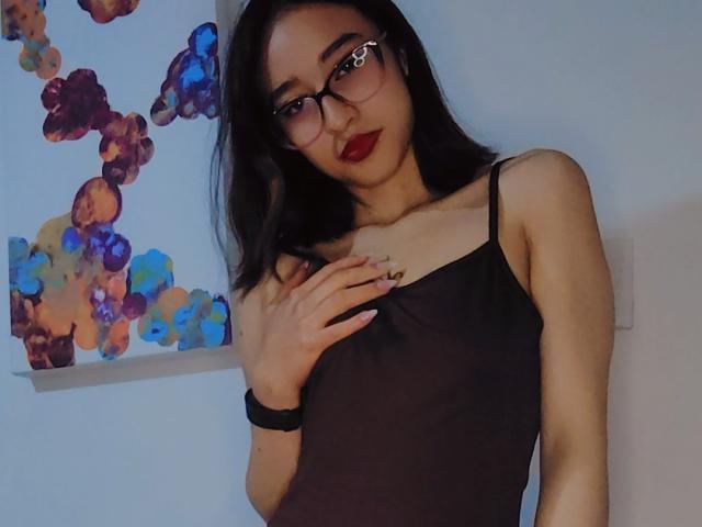 CamiVargas with dark hair, glasses, red lipstick, black spaghetti strap top, hand on chest, black wristband, medium tits, light skin, standing, blue wall, colorful abstract art.