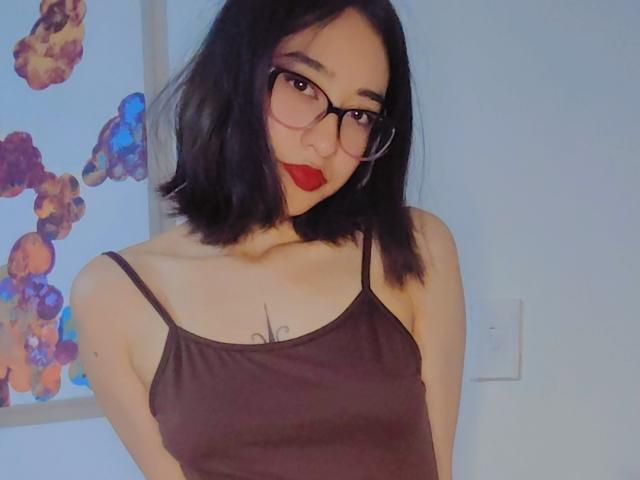 CamiVargas with black shoulder-length hair, red lipstick, and glasses, wearing a brown tank top, showing small tits and a tattoo below her collarbone, smiling slightly.