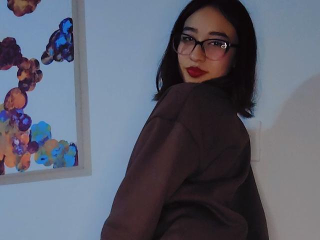 CamiVargas with dark brown bob, glasses, red lipstick, and black oversized shirt, looking over shoulder, showing her back and a small part of her ass. Painting in background.