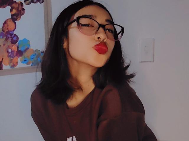 CamiVargas with dark hair, glasses, red lipstick, and a brown sweater, puckering her lips in a selfie, showing her nose ring.