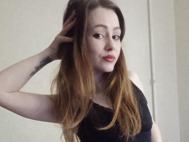 KendyOwers69 with long brown hair, wearing a black lace top, hand in hair, tattoo on forearm, pale skin, red lipstick, slight smile, looking at camera.