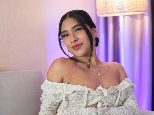 AntonellaPreschot has dark hair, a cute smile, and wears a white lace off-shoulder top, showing her tits and a pearl necklace. She's sitting on a couch with a lamp.