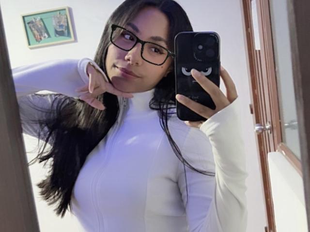 AnaRome with long black hair, glasses, and white zip-up, showing off her tits in a mirror selfie, making a peace sign with her right hand, holding a black phone.
