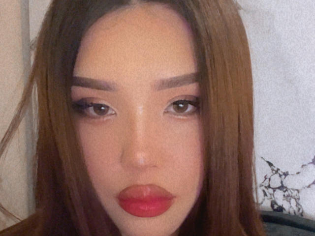 Close-up of MantyStyle with straight, medium-brown hair. She has light skin, prominent eyebrows, and brown eyes with long, thick eyelashes. Her lips are full and painted a bright red. The composition centers on her face, filling most of the frame. The texture of her hair is smooth, and her expression is neutral. The background is slightly blurred and contains a small, black-and-white illustration on the right.