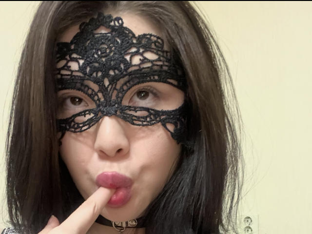 MantyStyle with black lace mask, puffy pink lips, finger in mouth, black choker, brown hair, pale skin, and a sexy, seductive expression. Her eyes are slightly open.