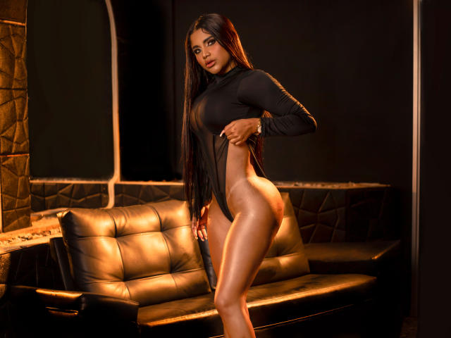 Shalimary with long black hair, wearing a sheer black top, exposing her bare ass and sideboob, standing in front of a black leather couch, under warm lighting.
