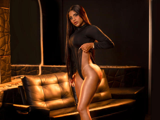 Shalimary, con el pelo largo y negro, vestida con una camiseta negra ajustada de manga larga, la baja para mostrar su trasero brillante y desnudo, y sus glúteos sin vello púbico, de pie en una habitación con poca luz..
