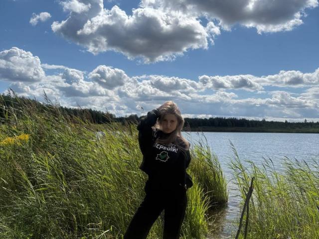 NiksaLove stands in tall green grass, wearing a black shirt with a white design, hands in her hair, looking serious, near a lake with a cloudy blue sky.