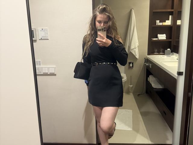 NiksaLove, con cabello rubio ondulado, vestido negro de manga larga, cinturón y zapatos de tacón marrón, sosteniendo un teléfono en un selfie frente al espejo, con un bolso negro en el brazo derecho, de pie en el baño..