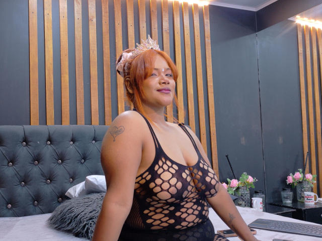 AliceCox, a plus-sized woman with light brown skin and red hair, wears a black fishnet dress that reveals her large breasts and tattoos on her right shoulder and upper arm. She has a pink floral headband and a confident smile. She's sitting on a bed with a tufted dark green headboard and wooden slats in the background. The room includes pink flowers and a laptop on a table. The lighting is soft, highlighting her features. She looks directly at the camera.