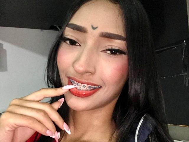 AdelaBanks with black hair, red lipstick, and a small crescent moon tattoo above her eyebrows, sucking on a white and pink lollipop with her right finger.