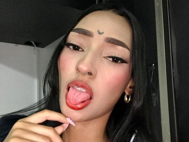 AdelaBanks has long black hair, heavy dark eyebrows, and is sticking out her tongue with red lipstick on it. She has a small crescent tattoo above her eyebrows, pink nail polish, and is holding her tongue with her left hand. She wears gold hoop earrings. Her eyes are lined with black eyeliner, and she has a slight smile. The lighting is even, and the focus is on her face. The image is a close-up selfie.