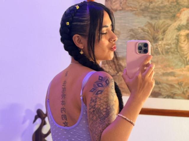 LunaDesire with braided hair, holding phone, wearing a purple tank top, showing tattoos on back and arm, taking a selfie, pink phone, pink lipstick, bracelet, nose ring.