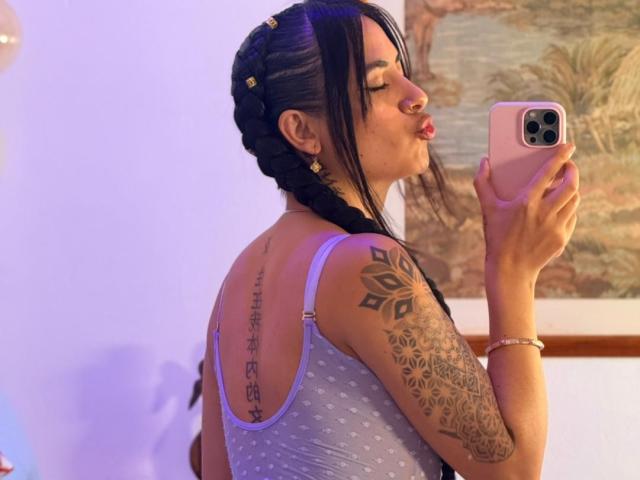 LunaDesire, with braided black hair, holding a pink phone, kissing reflection, tattooed arm, grey polka-dot top, back tattoo, wearing gold earrings, purple wrist bracelet.