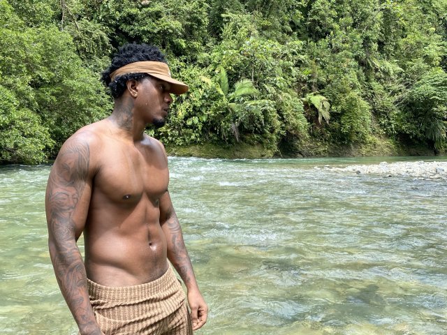 LoganDrakex stands in a clear river, shirtless with a tan visor and brown, ribbed shorts. He has dark curly hair, a tattooed right arm, and visible chest tattoos. His skin is medium brown, and he has a muscular, toned build. The river's clear water and rocky shore contrast with the dense, green forest in the background. The composition focuses on LoganDrakex's upper body, emphasizing his tattoos and muscular physique. The natural light highlights his defined muscles and tattoos.
