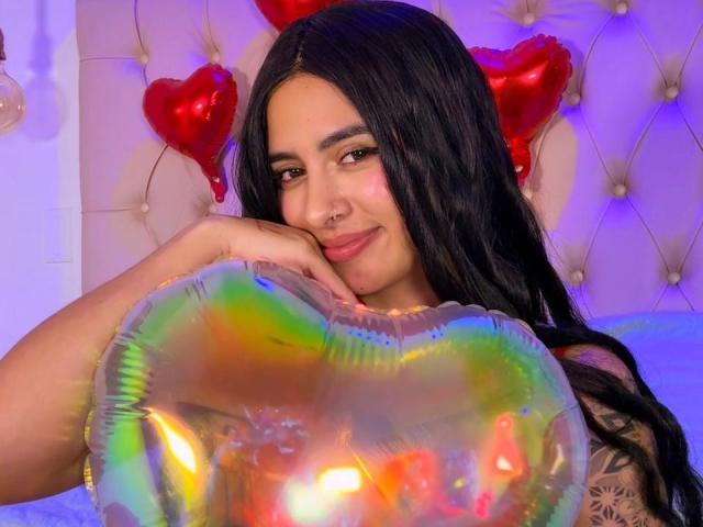 LunaDesire with long black hair, nose ring, and tattoo, holding a shiny rainbow heart balloon, smiling at the camera. Red heart balloons in the background.