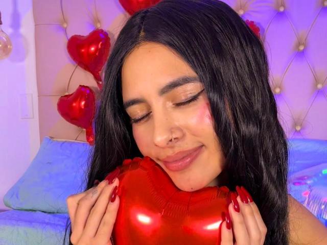 LunaDesire with long black hair, closed eyes, and red nail polish holding a shiny red heart, smiling softly. She has a nose ring and is in a pink-tufted room.
