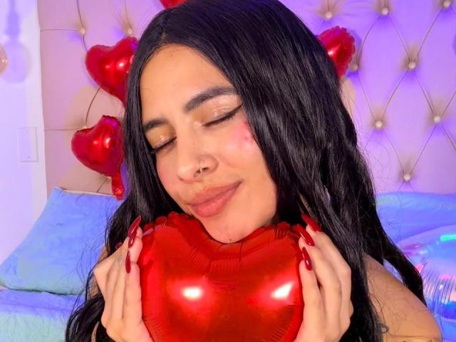 LunaDesire with long black hair, closed eyes, nose ring, and red lipstick, holding shiny red heart, red nails, pink blush, and smiling softly. Red heart balloons in background.